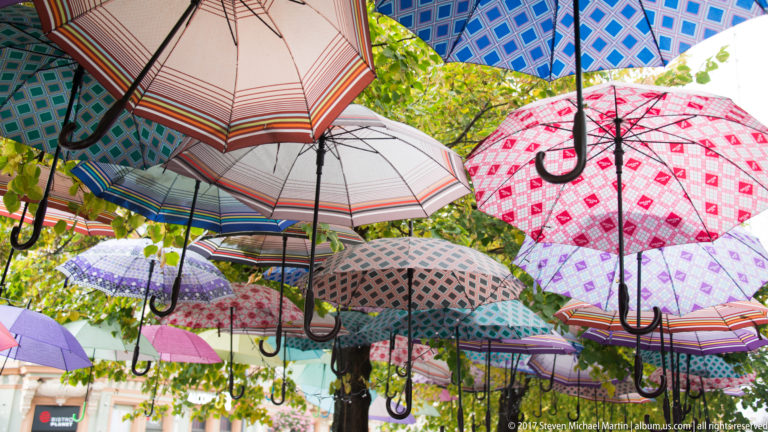 Colorful patterned umbrellas suspended overhead among green tree foliage in a pedestrian area.