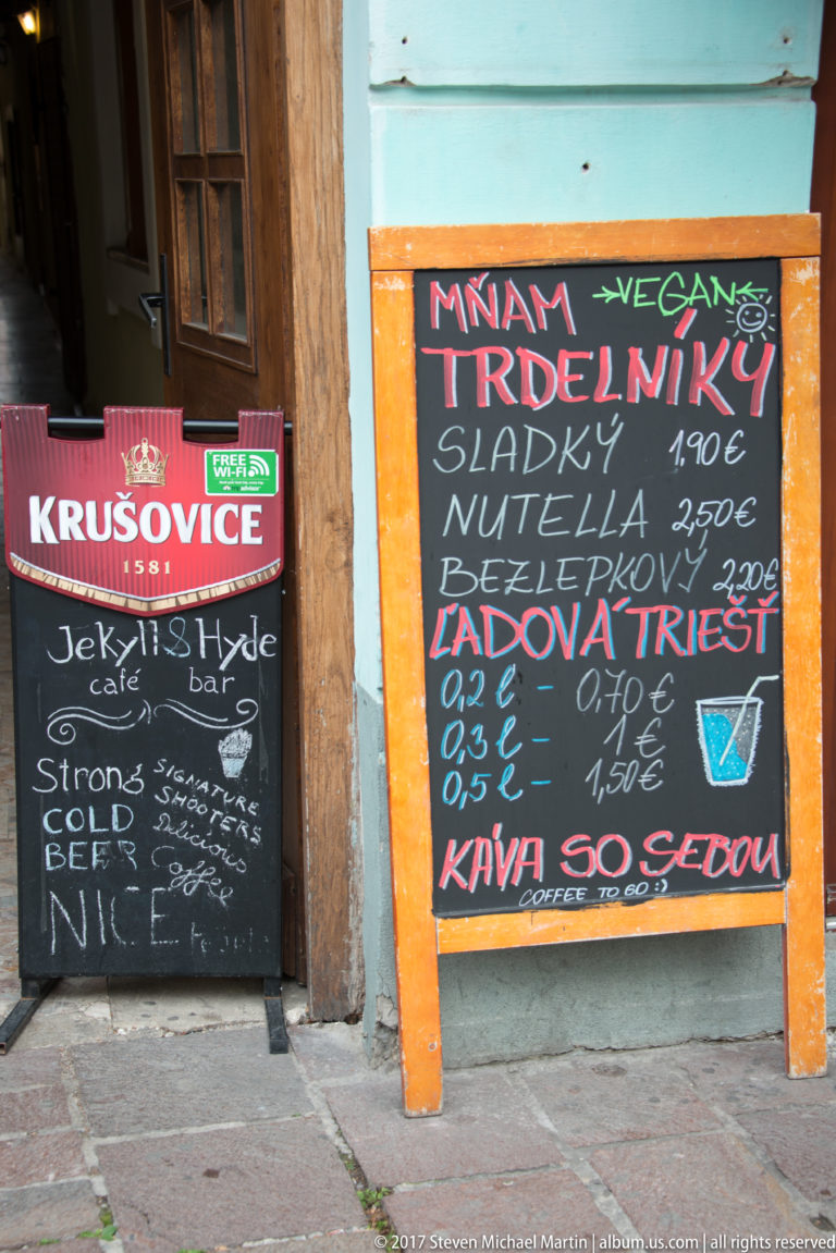 Handwritten café chalkboards in Slovak advertising trdelník, drinks, and euro prices beside a doorway.