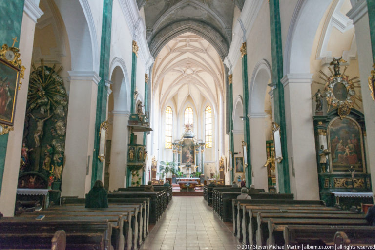 Long Gothic church nave with ribbed vaults, wooden pews, side altars, and a bright sanctuary at the far end.
