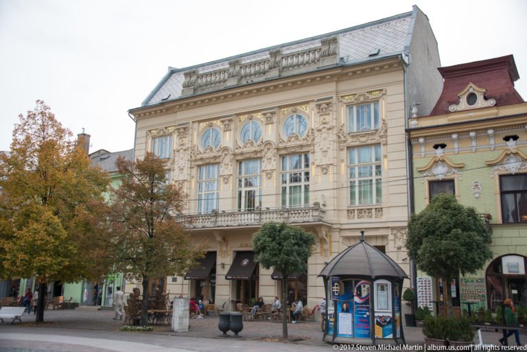 Ornate pale historic building with tall decorated windows, central balcony, café frontage, trees, and a kiosk.