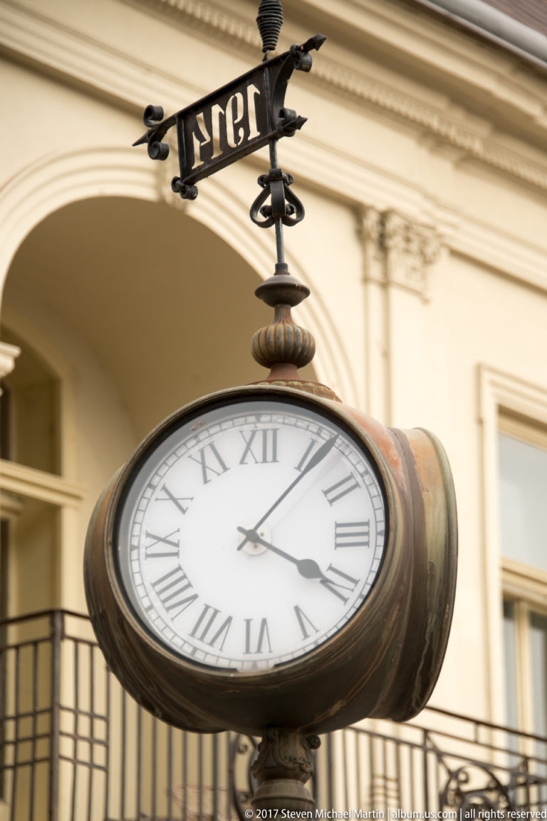Round ornamental street clock with Roman numerals, dark metal casing, finial, and small weather vane above.