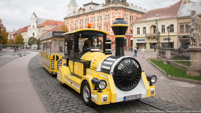 Yellow sightseeing road train with locomotive-style front traveling through a historic square in Košice.
