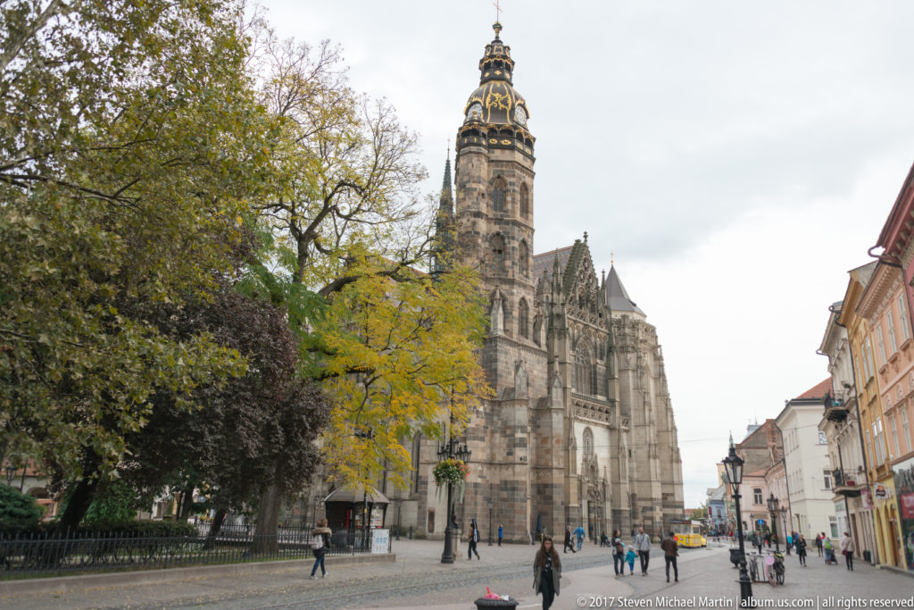 St. Elisabeth Cathedral rising beside a pedestrian square, with tall tower, Gothic stonework, trees, and people.