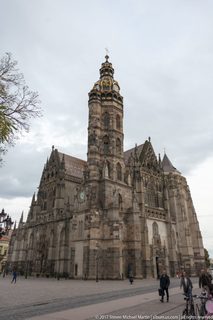 Broad side view of St. Elisabeth Cathedral with massive tower, buttresses, Gothic windows, tiled roofs, and open plaza.