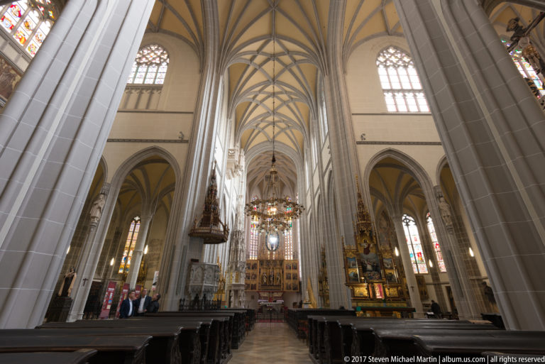 Angled cathedral nave view with high ribbed vaults, massive piers, hanging chandelier, stained glass, and side altarpieces.
