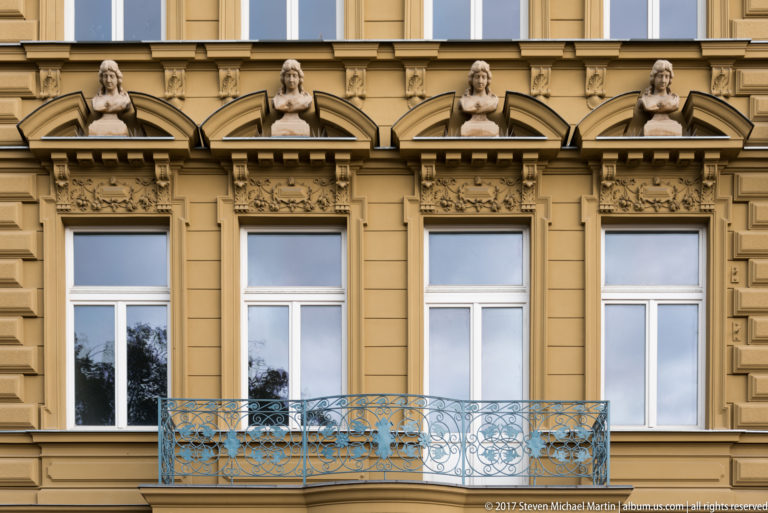 Four historic window bays with sculpted female busts, ornate stucco decoration, and a blue wrought-metal balcony.