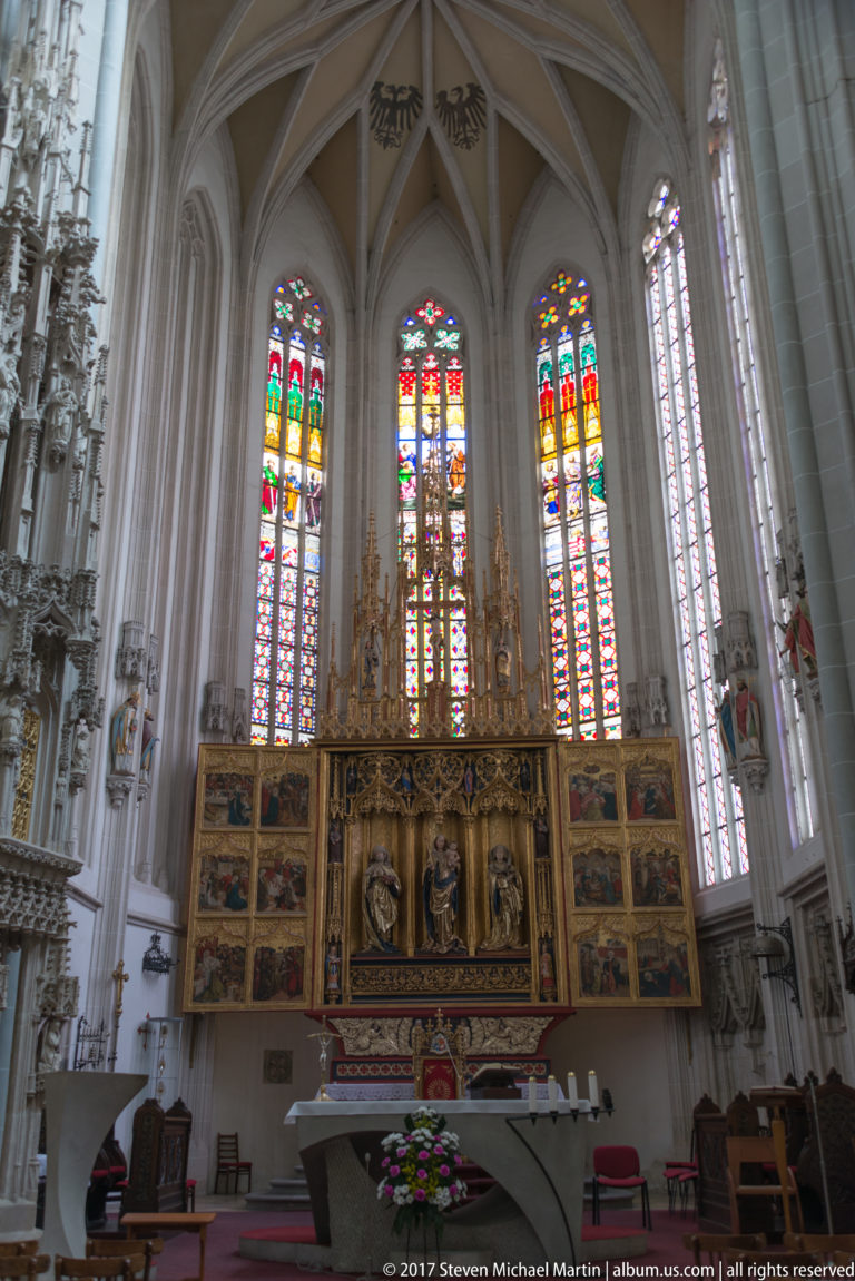 Gothic cathedral sanctuary with winged altarpiece, sculpted figures, painted panels, and tall stained-glass windows.