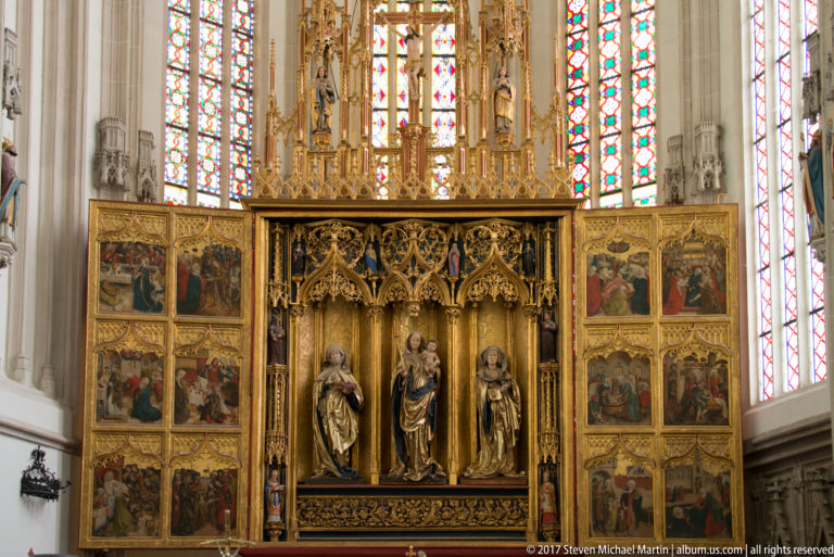 Open Gothic winged altarpiece with three sculpted figures, painted side panels, gilded tracery, and stained glass behind.