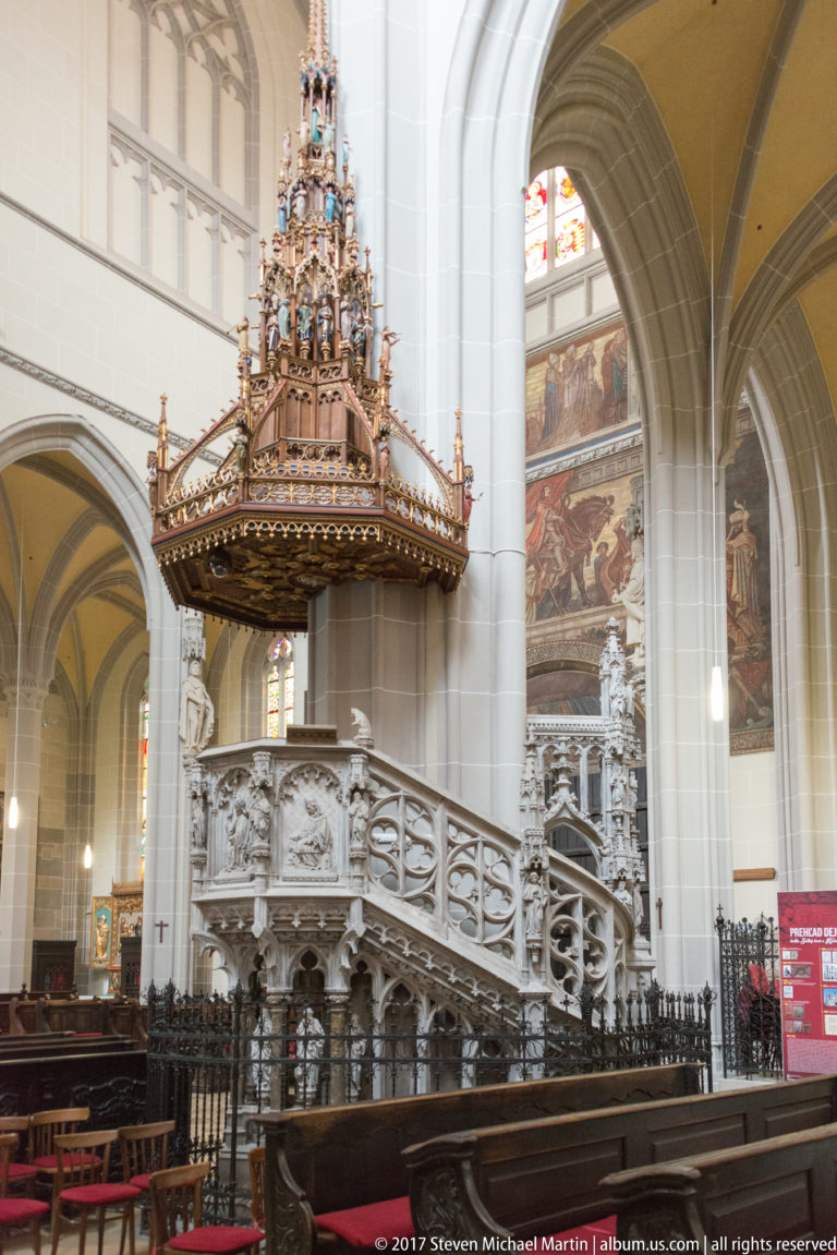 Ornate cathedral pulpit with carved stair, openwork balustrade, relief sculpture, and tall spired canopy.