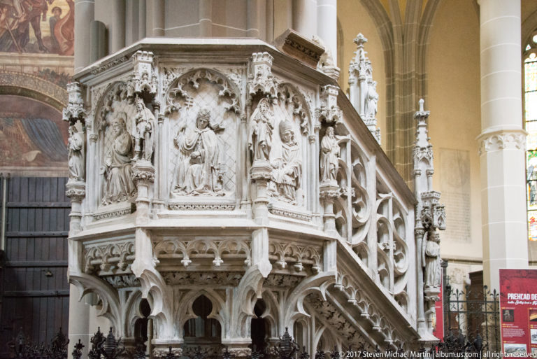 Close view of a pale stone pulpit with carved figures, pointed arches, tracery, quatrefoils, and an ornate stair.