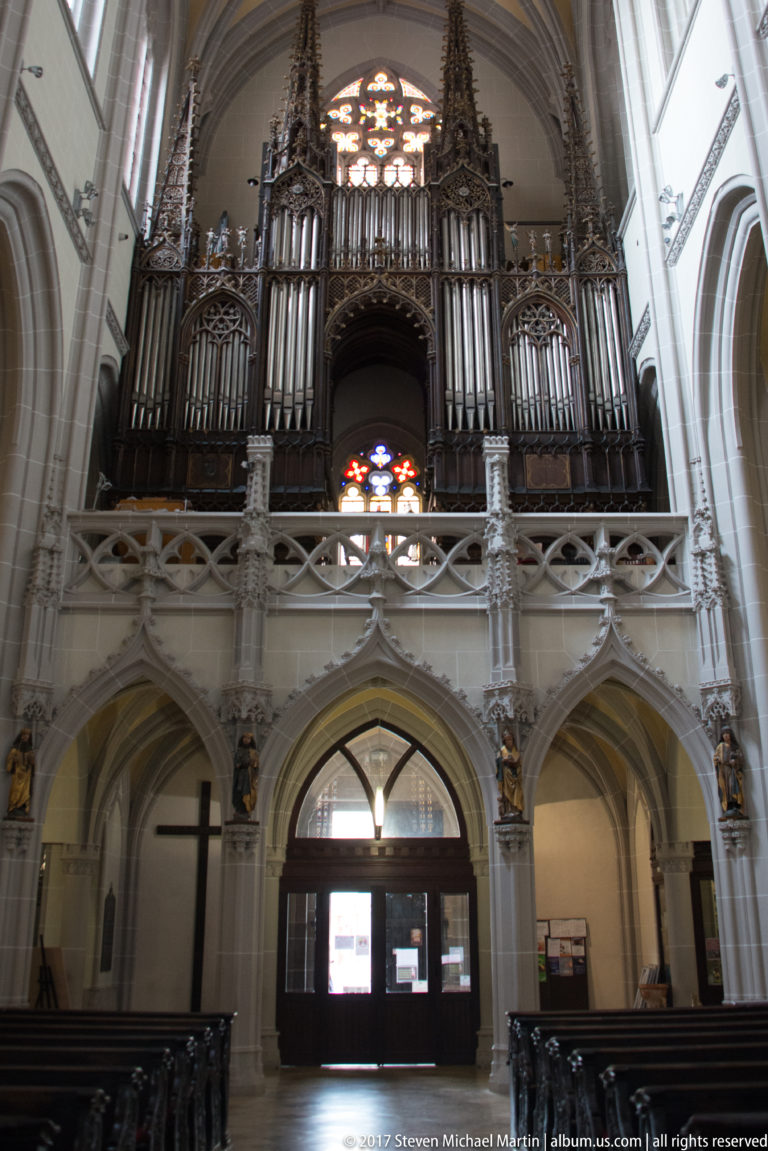 Large cathedral pipe organ on a stone gallery with metal pipes, Gothic woodwork, and stained glass above the entrance.