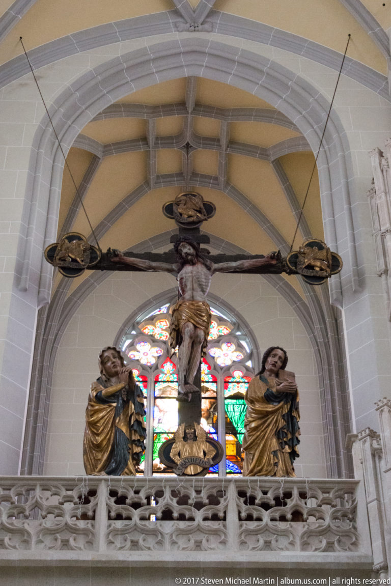 Large crucifix with two flanking sacred figures above a stone balustrade, framed by stained glass and ribbed vaults.