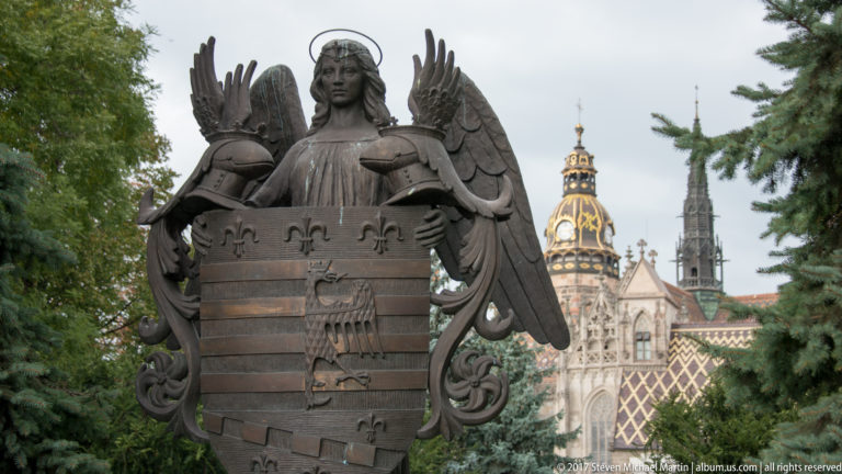 Bronze winged angel holding a heraldic shield, with St. Elisabeth Cathedral blurred among trees behind.
