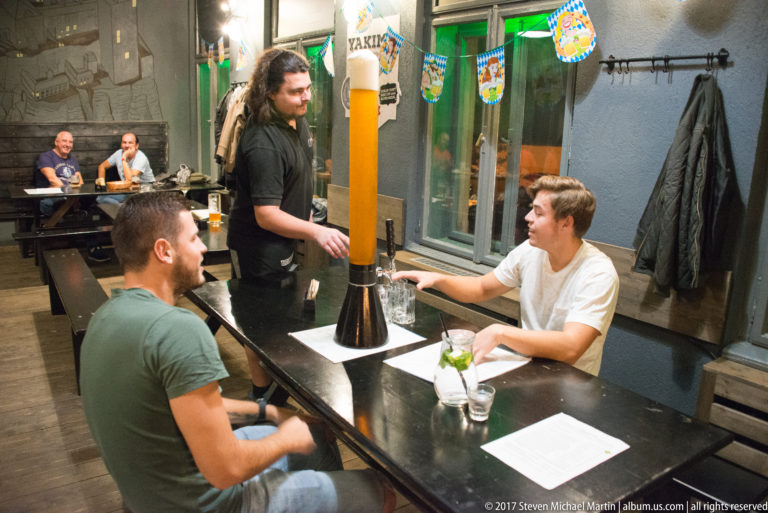 Three men around a table with a tall transparent beer tower in a casual Košice beer hall.