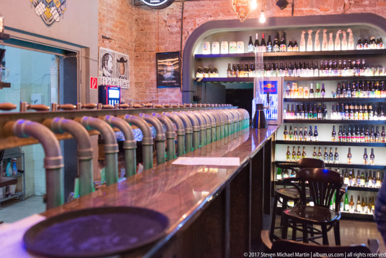 Long bar lined with many metal beer taps, backed by shelves filled with bottles in a brick-walled interior.