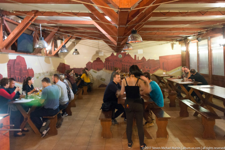 Busy beer hall with long wooden tables, benches, exposed roof trusses, pendant lights, and a city mural.