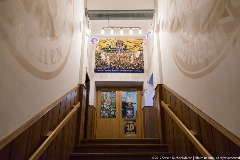 Narrow stairway descending between wood-paneled walls to a sticker-covered doorway in a bar interior.