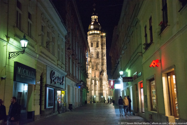 Narrow nighttime street with historic façades and pedestrians leading toward an illuminated Gothic cathedral tower.