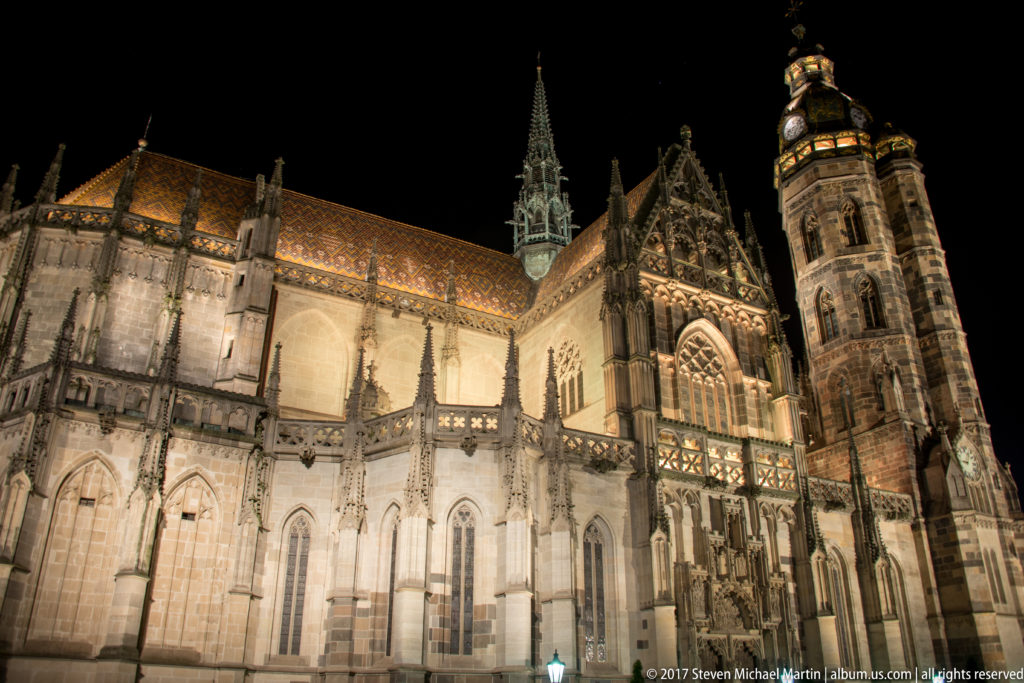 Floodlit St. Elisabeth Cathedral at night with Gothic buttresses, traceried windows, patterned roof, gables, and tower.