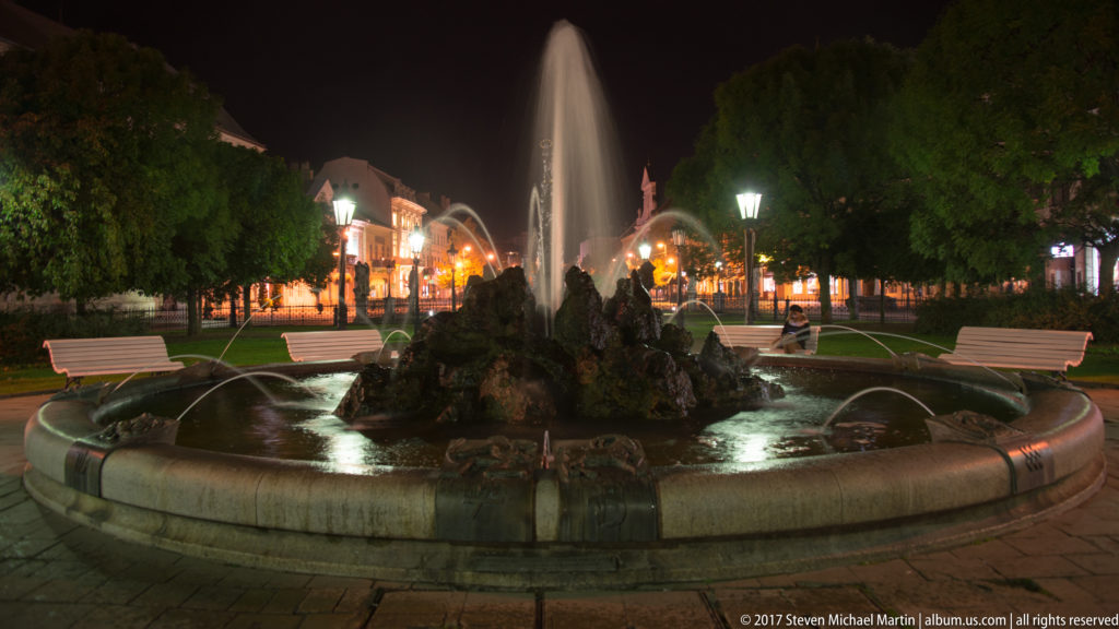 Circular stone fountain at night with tall central jet, rockwork, smaller water arcs, benches, trees, and lamps.