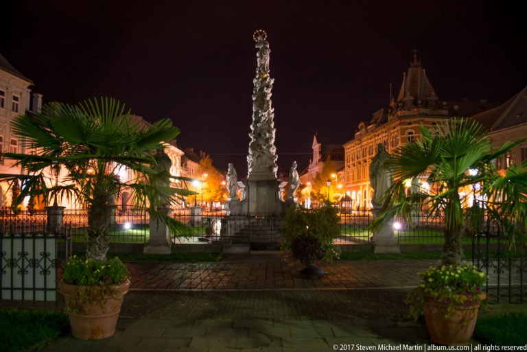 Illuminated Immaculata plague column with Virgin Mary figure, base sculptures, planters, and historic buildings at night.