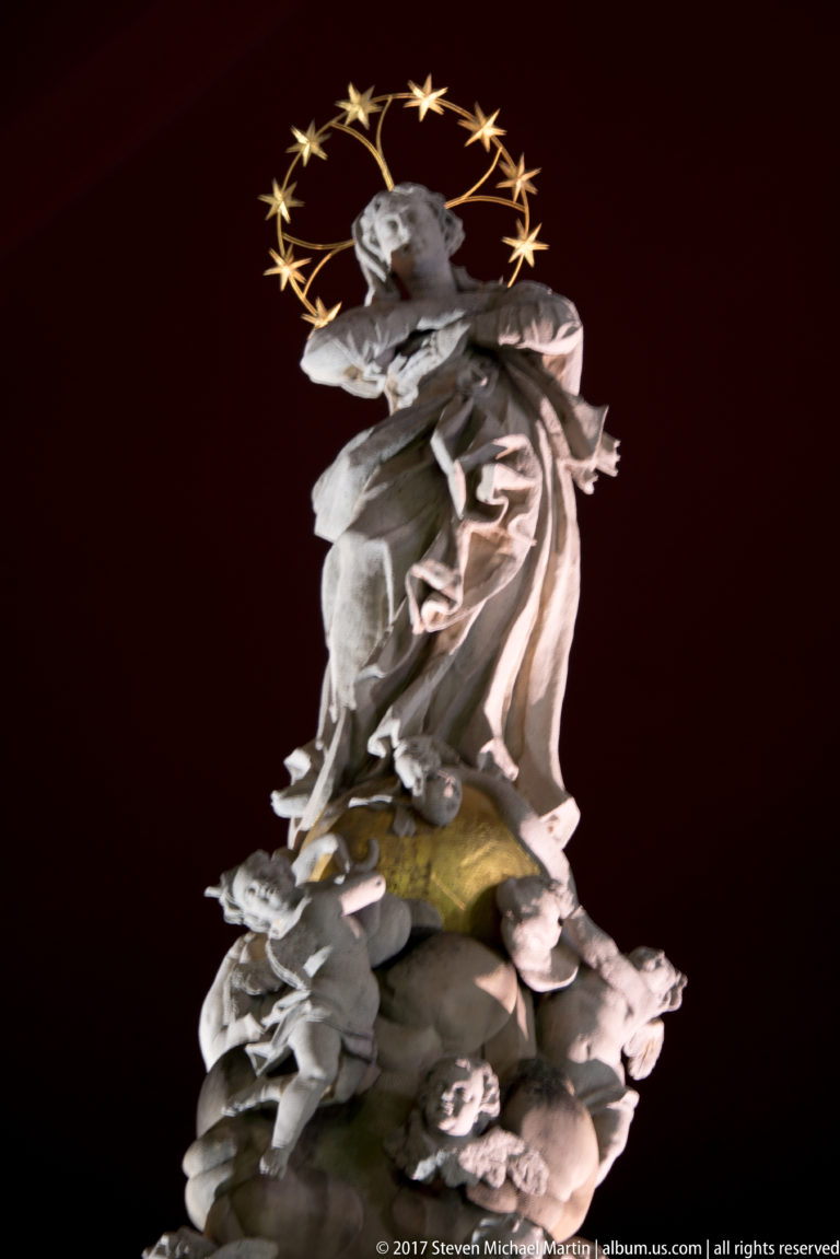 Illuminated Virgin Mary figure above clouds and angels, surrounded by a halo of stars against the night sky.
