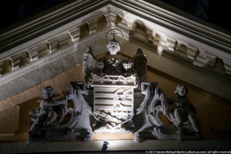 Illuminated heraldic shield with crest, sculpted supporters, and scrolling ornament on a historic building corner.