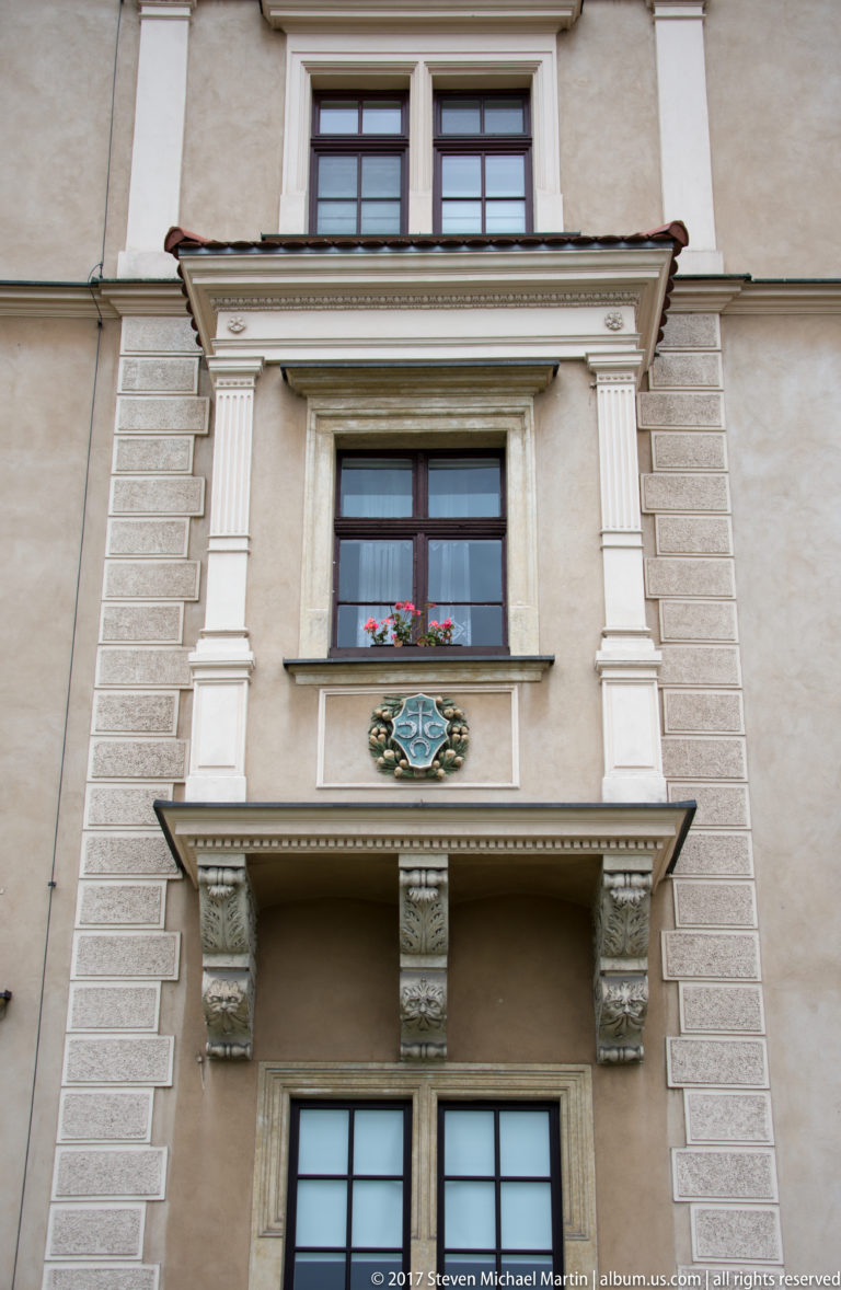 Two stacked windows with carved surrounds, projecting hoods, brackets, and an ornamental crest on a pale façade.