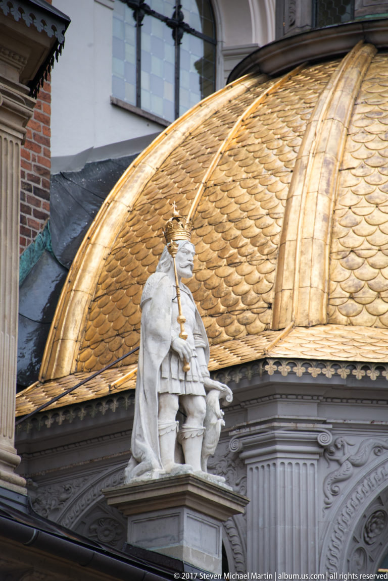Stone royal figure holding a staff in front of a patterned gilded dome at Wawel Cathedral.