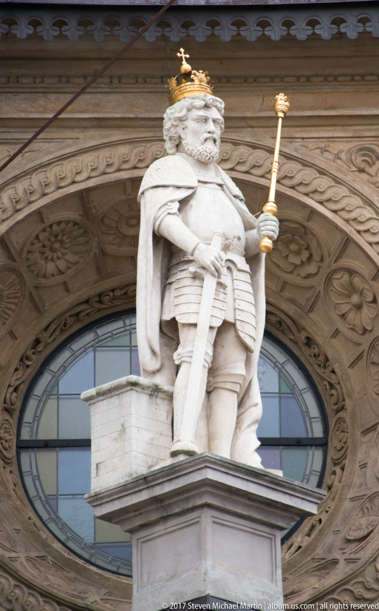 Crowned stone figure in armor holding a scepter before an ornate arched window and carved stone surround.