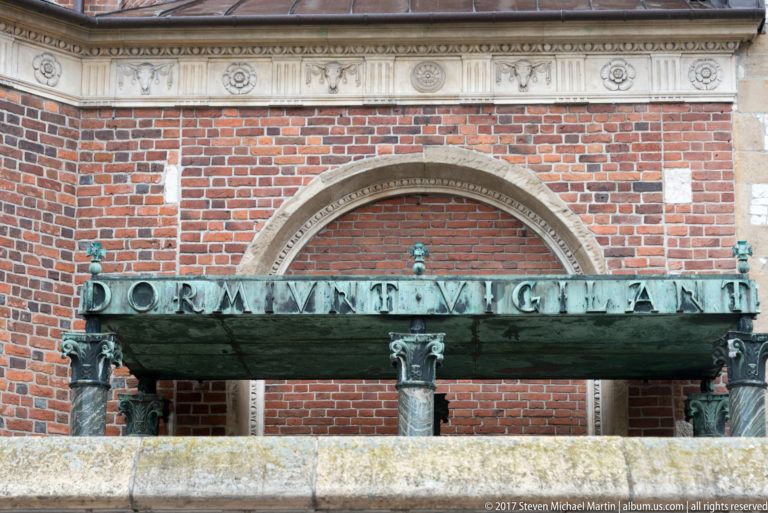 Green-patinated inscribed metal beam on decorative supports in front of a pointed brick arch.