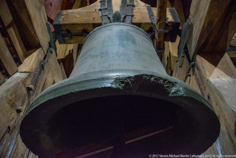 Large bronze bell seen from below, suspended from heavy wooden beams and iron fittings inside a bell tower.