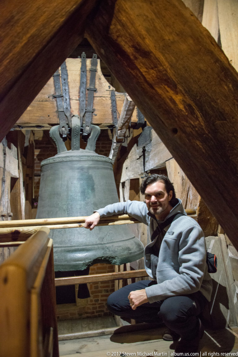 Visitor crouching beside a large bronze bell amid heavy timber beams and iron suspension hardware.