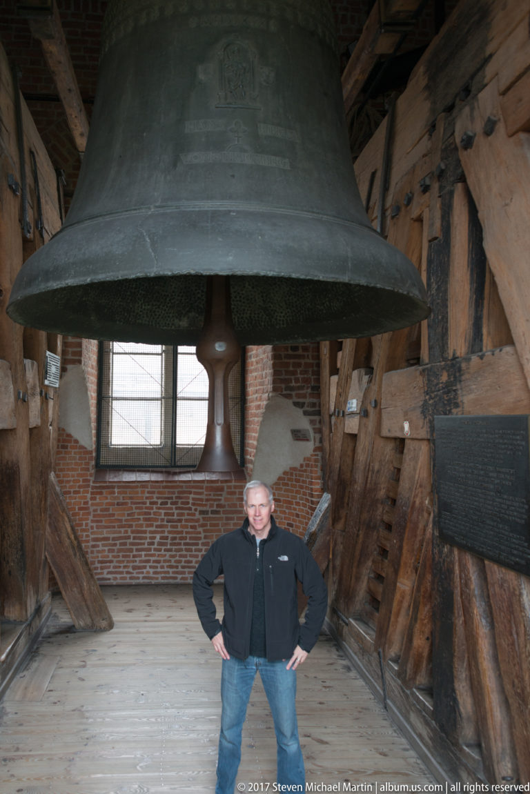 Large suspended bell towering over a visitor inside a timber-framed bell chamber with brick walls.