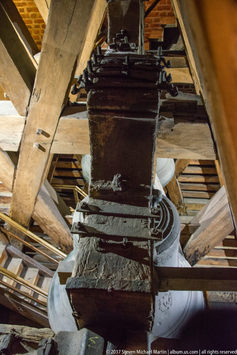 Close overhead view of a heavy wooden bell headstock wrapped with iron bands above part of a bell.