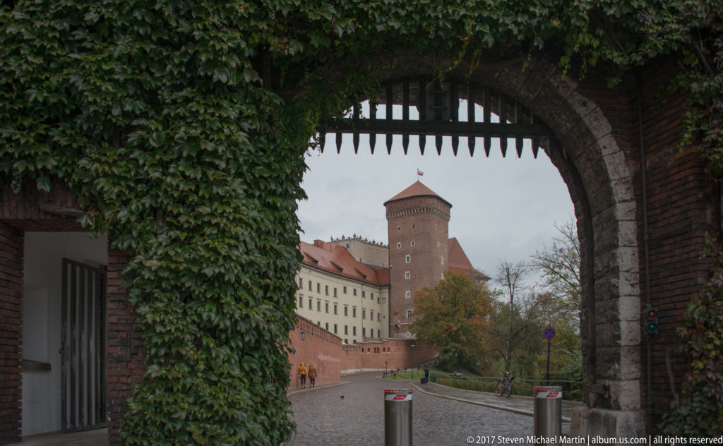 Ivy-covered arched gateway framing a cobbled road, brick tower, and palace buildings inside Wawel Castle.