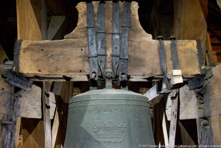 Inscribed bronze bell hanging beneath a large wooden headstock reinforced with vertical iron straps.