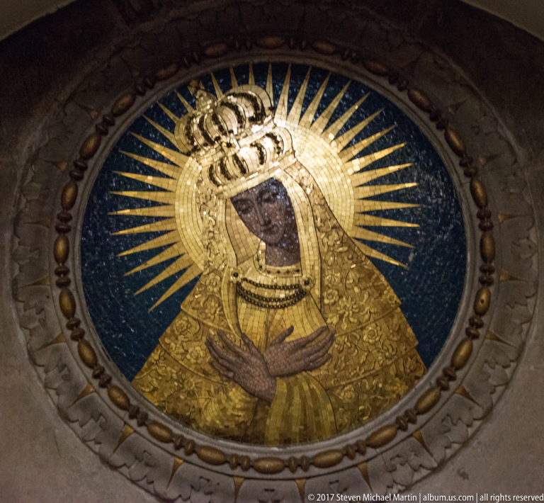 Circular mosaic of the crowned Virgin Mary with crossed hands, gold robes, and radiating halo on a blue ground.