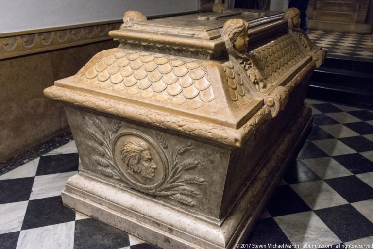 Carved stone sarcophagus with a portrait medallion, scale-patterned sides, and sculpted corner heads on a tiled floor.