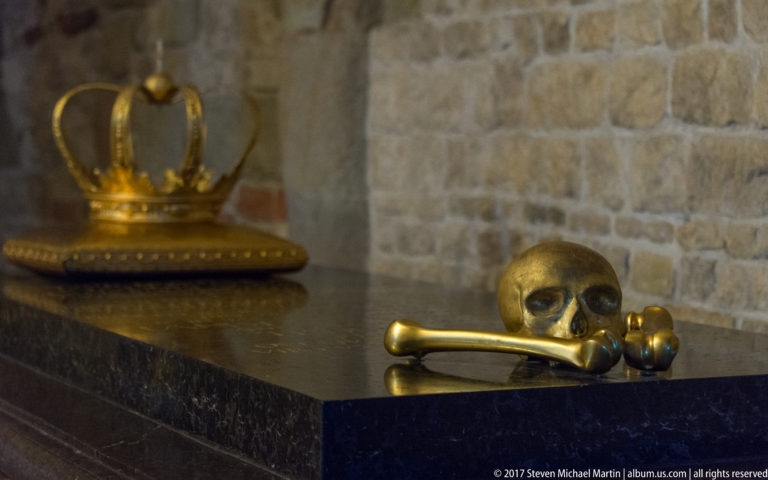 Gilded skull and crossed bones on a dark ledge, with a ceremonial crown blurred in the background.