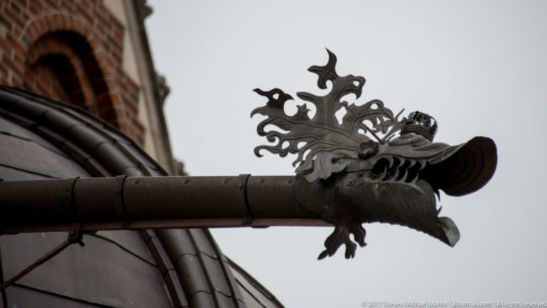 Dark metal rain spout ending in an ornate open-mouthed dragon head projecting from a building.
