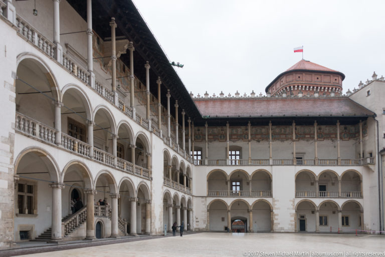Wide view of Wawel Royal Castle's arcaded courtyard, with multi-level galleries and a brick tower flying the Polish flag.