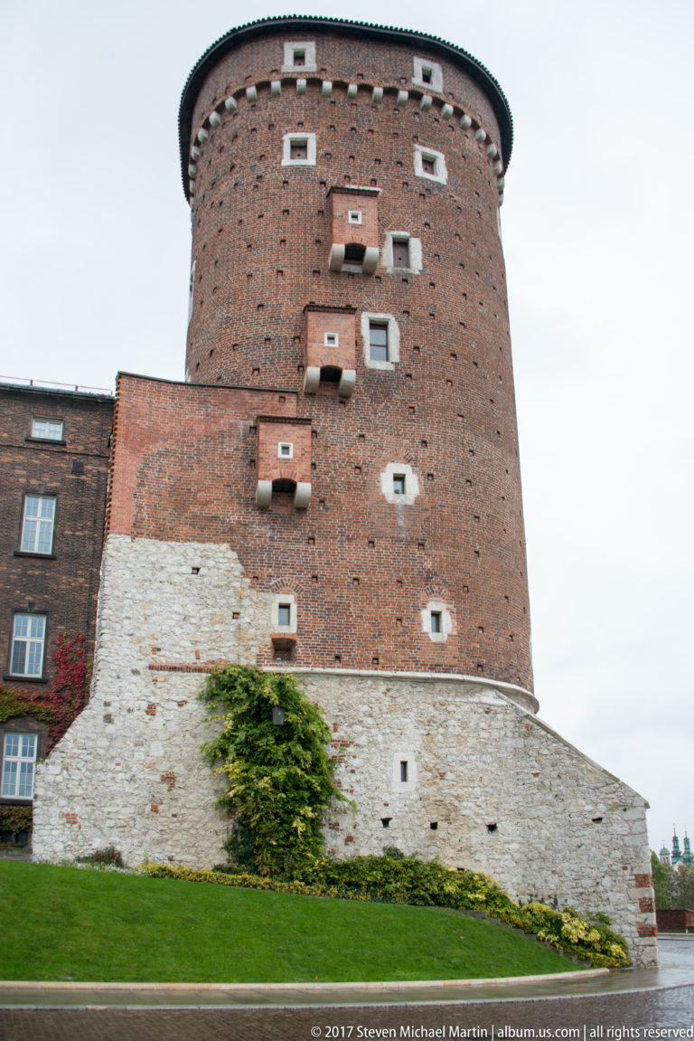 Cylindrical red-brick defensive tower rising above a sloped pale-stone wall and green lawn.