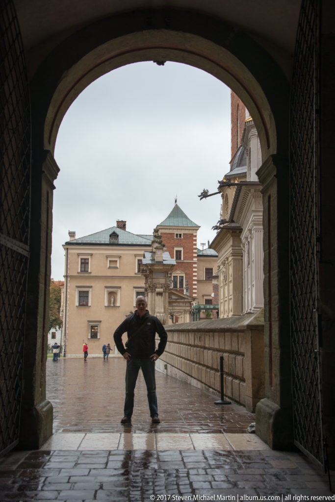 Visitor standing in silhouette beneath a dark arch, looking into the wet courtyard and palace buildings beyond.