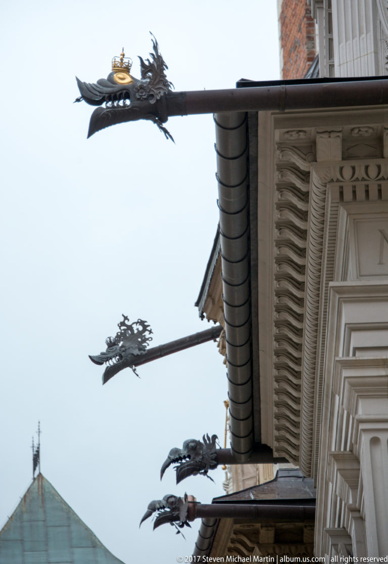 Multiple dark metal dragon-headed rain spouts projecting from a roofline above carved stone cornices.