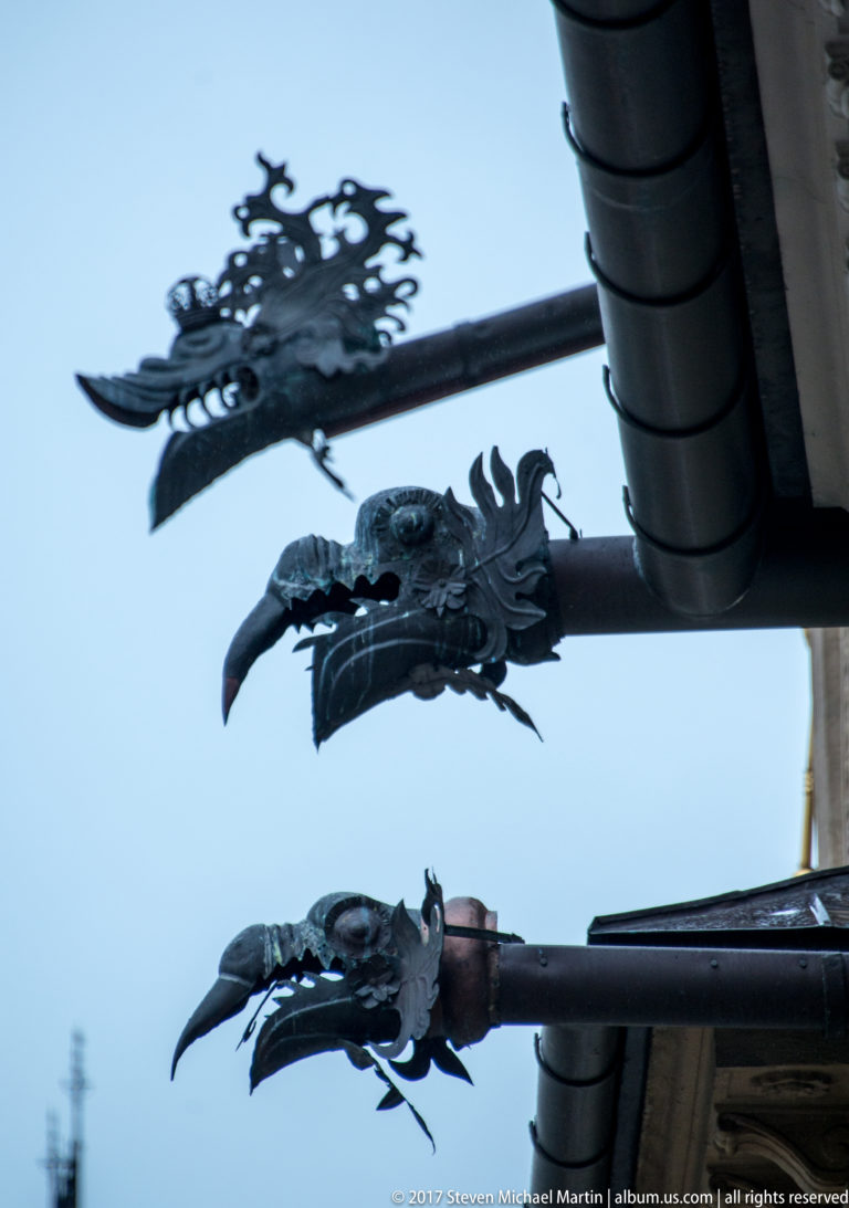 Three dark metal dragon-headed spouts with open jaws project outward against a pale blue-gray sky.