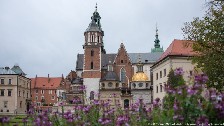 Purple flowers in the foreground with Wawel Cathedral's brick tower, chapels, green spires, and gold dome beyond.