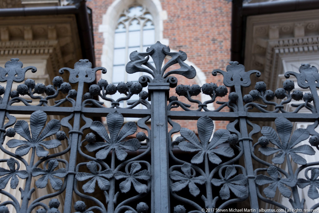 Ornate dark iron gate with symmetrical leaves, scrolls, floral forms, and heraldic motifs before a brick building.
