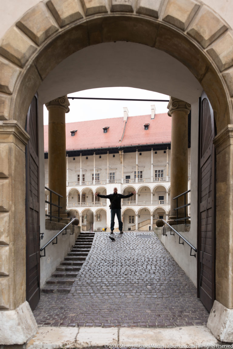 Stone arch and open wooden doors frame a cobblestone ramp leading to Wawel Castle's arcaded courtyard.