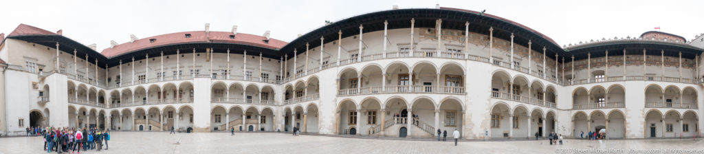 Wide view of Wawel Royal Castle's white multi-story arcades curving around a broad paved courtyard.